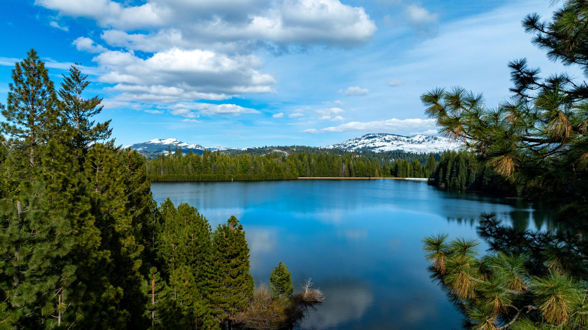 Les nuages ​​planent au-dessus d'un lac bleu vif entouré de conifères.  Des montagnes enneigées s'élèvent en arrière-plan.