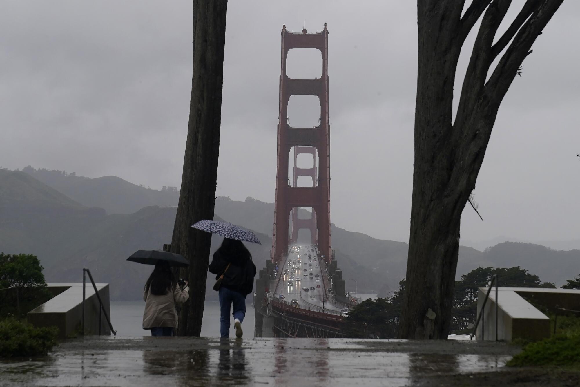 Deux personnes tiennent des parapluies pendant qu'elles marchent sous la pluie.  Le Golden Gate Bridge s'élève en arrière-plan.