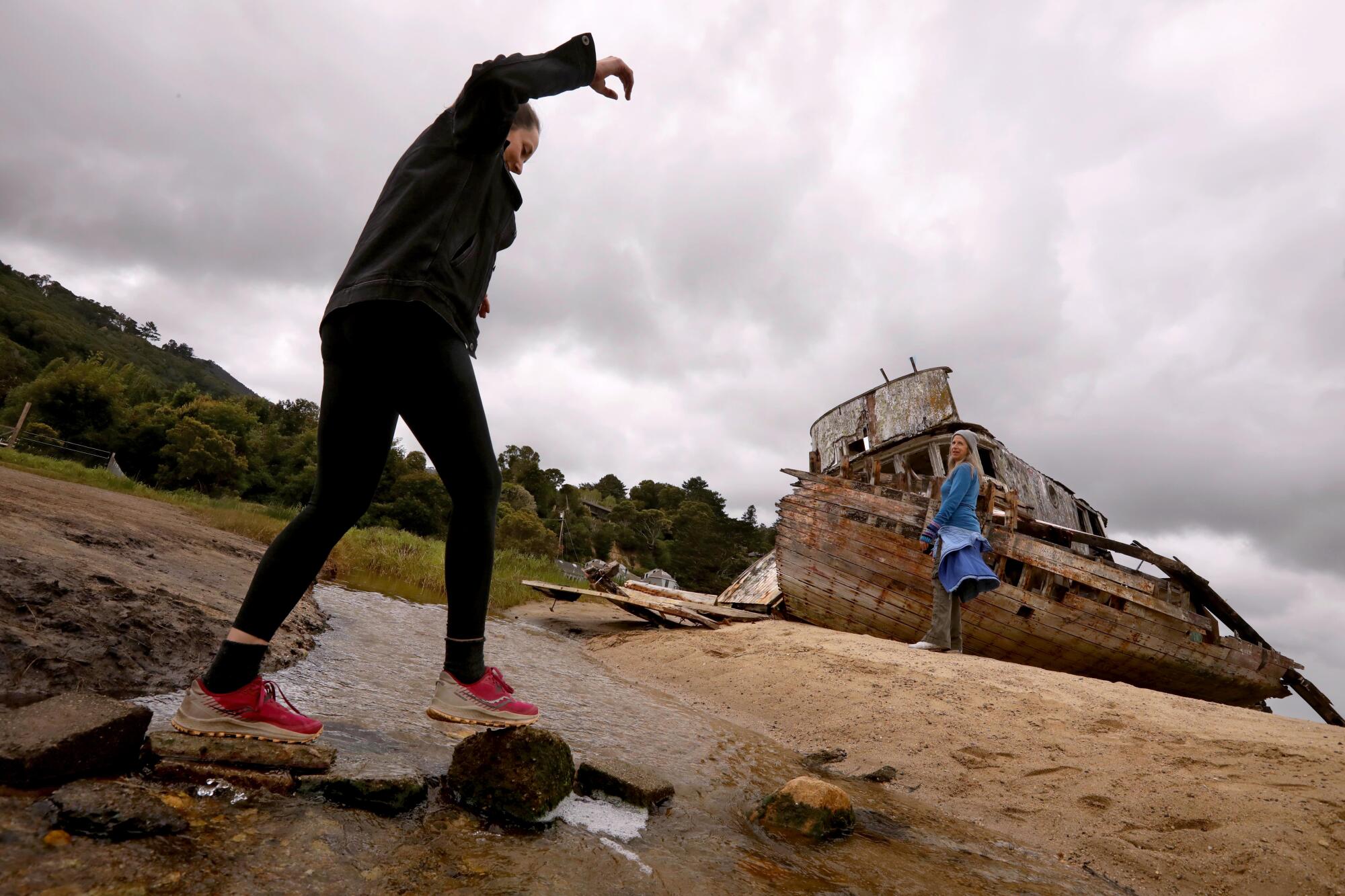 Une femme traverse un rivage rocheux près d’un bateau de pêche abandonné.