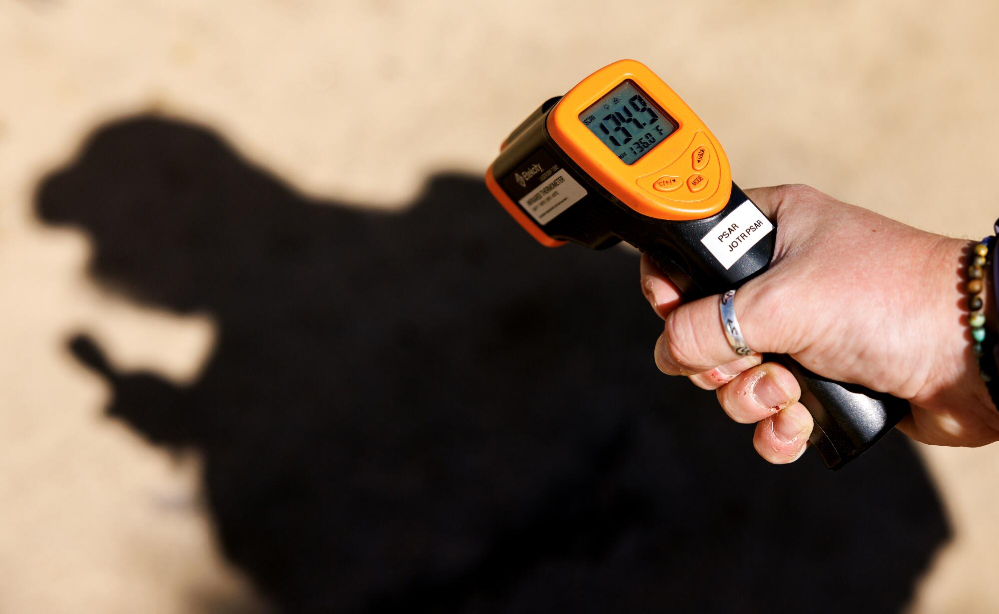 Une main pointe un thermomètre vers le sol du désert. Un indicateur numérique indique 134,9 degrés. 