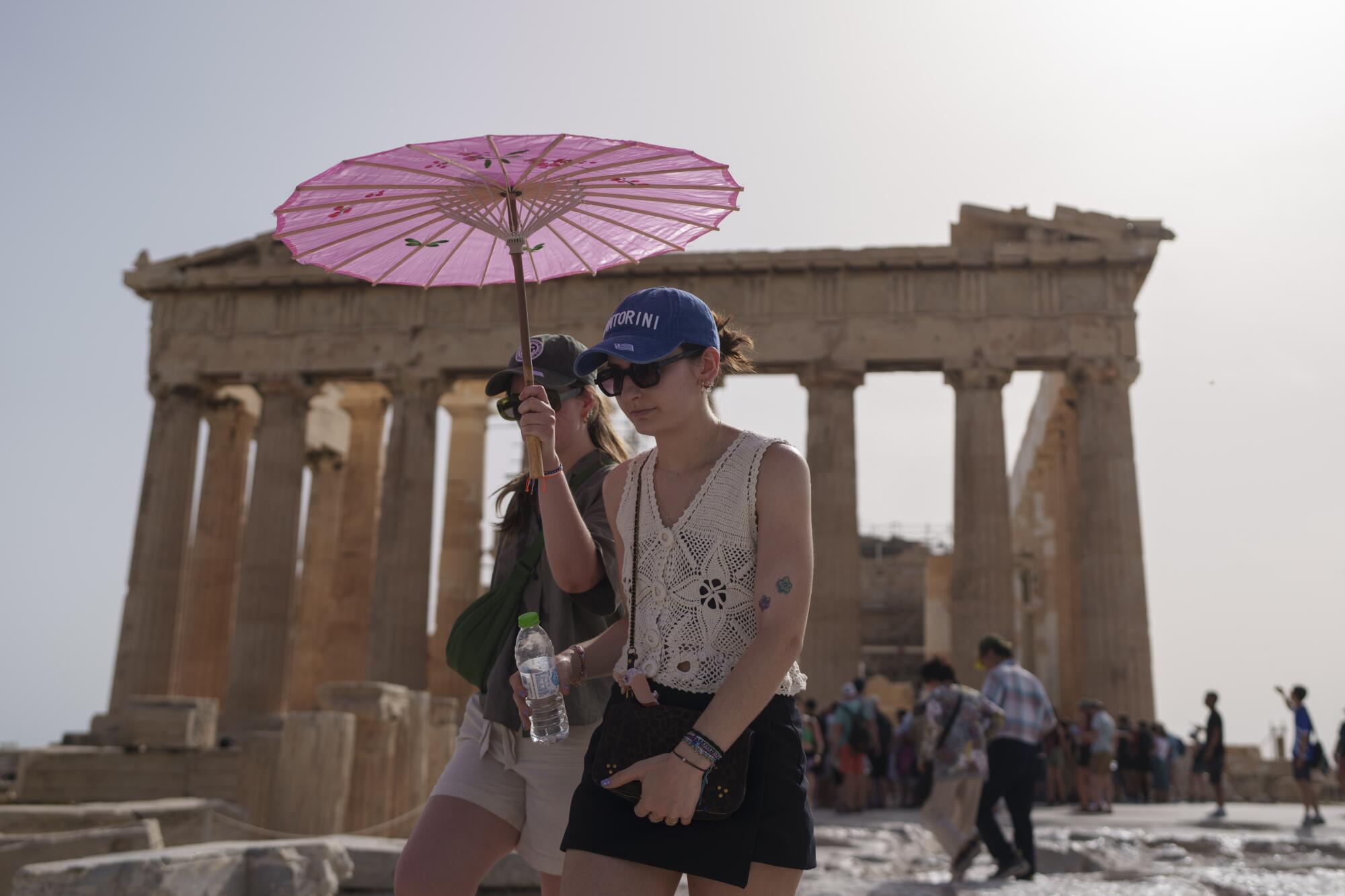 Deux femmes marchent sous un parasol rose tandis que le Parthénon se profile à l'arrière-plan.