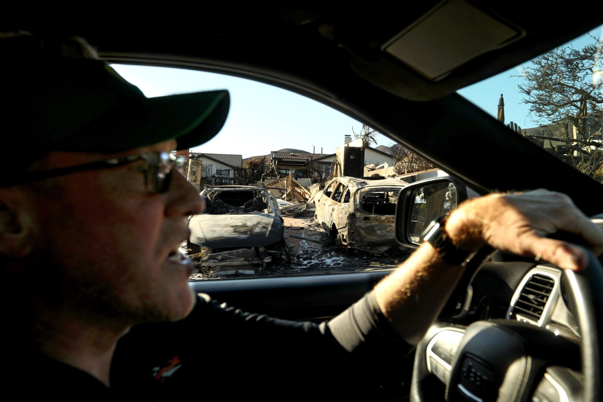 Un homme au volant d’une voiture passe devant des maisons et des véhicules incendiés.
