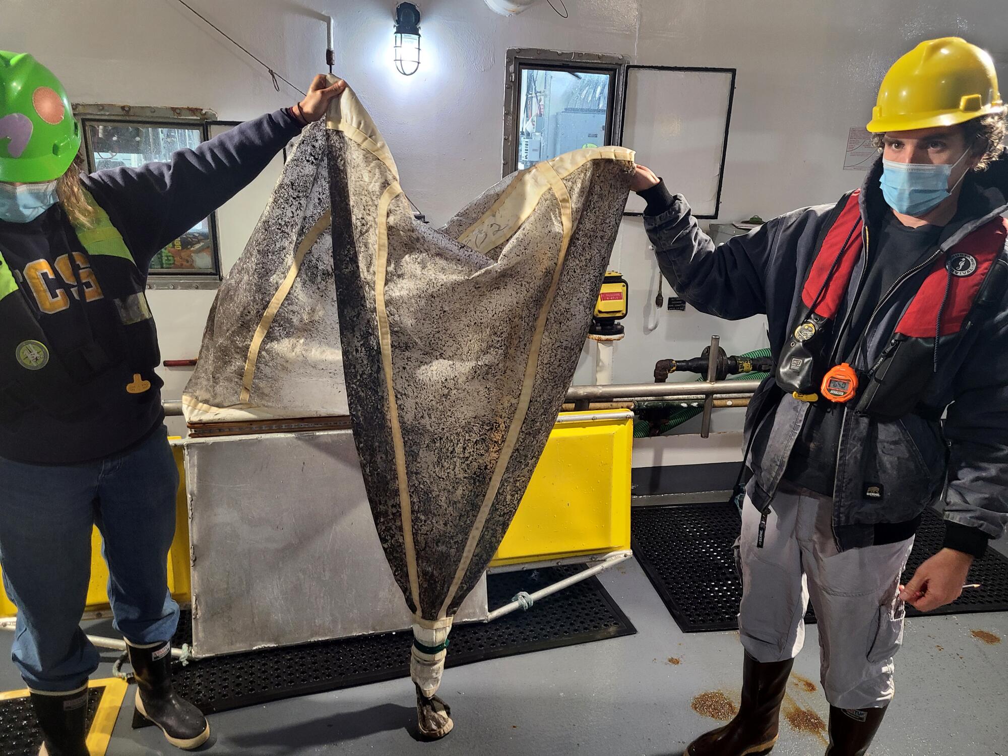 Deux hommes portant des respirateurs et des casques tiennent entre eux un filet en forme d'entonnoir obstrué avec des débris noirs