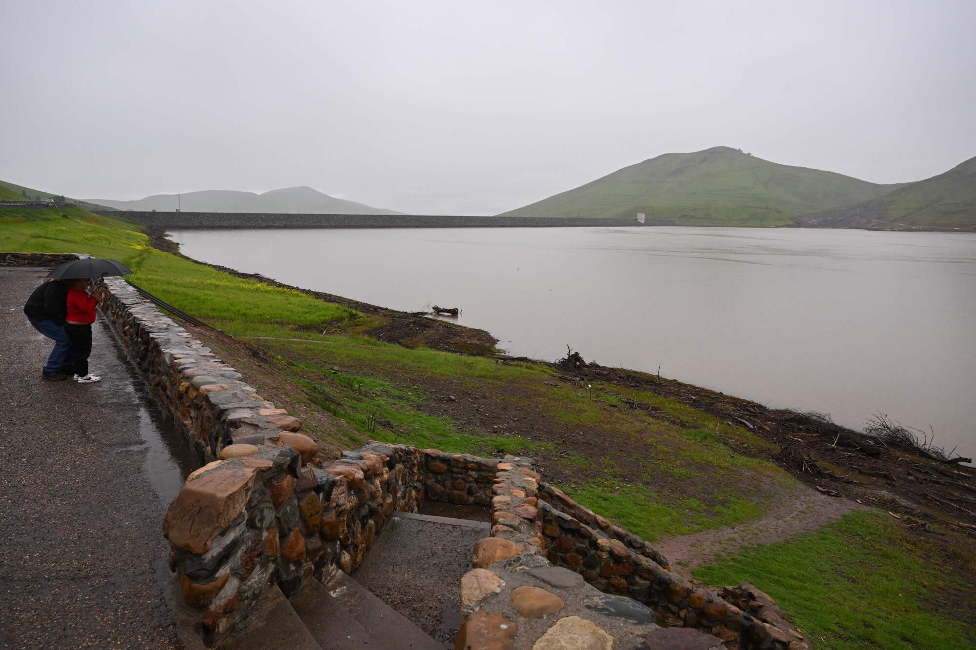 Les gens derrière un mur de pierre basse regardent une piscine complète d'eau derrière le barrage de Schafer formant le succès du lac.