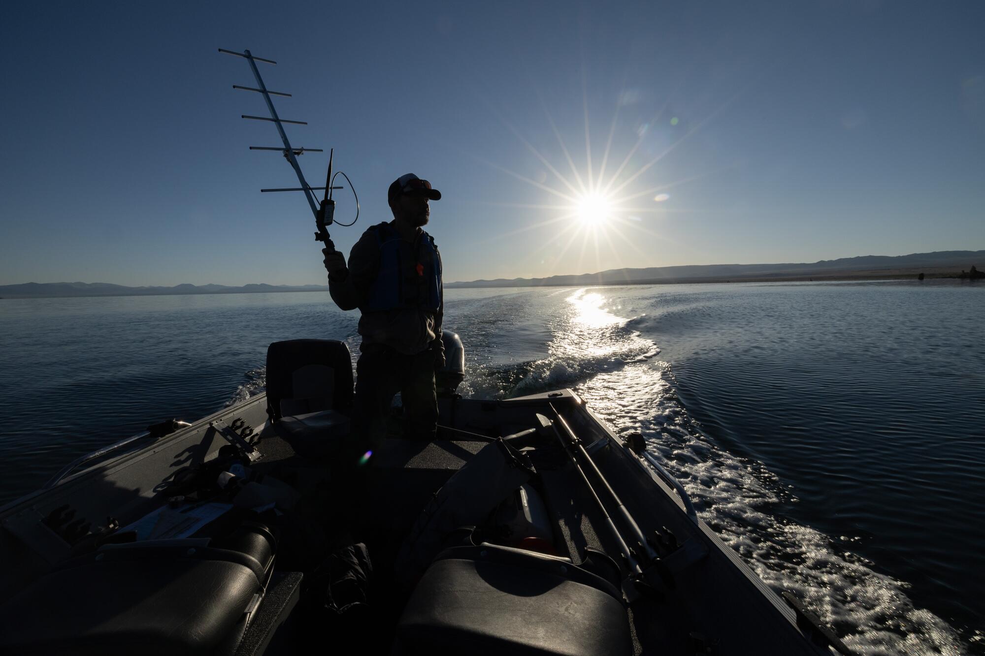 Un homme dans une casquette, vu dans l'ombre, tient une antenne dans un bateau sur un lac, avec le soleil bas à l'horizon