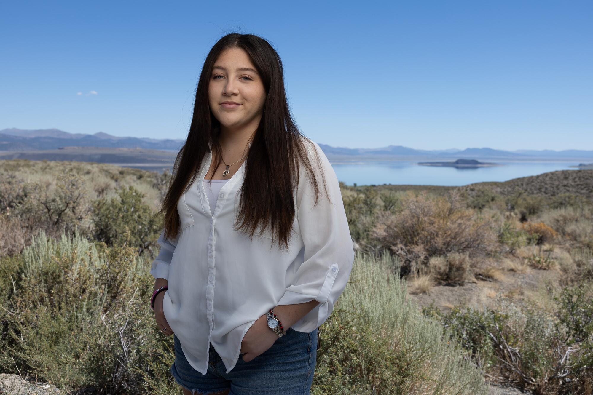 Une jeune femme aux longs cheveux noirs, dans une chemise et un jean à manches longues blanches, debout au milieu des arbustes près d'un lac