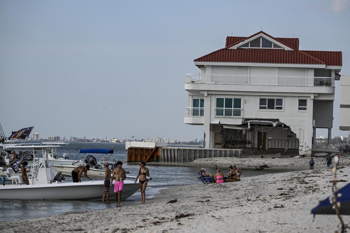 Les gens sont assis sur une plage alors qu'ils assistent à une défilé de bateau près d'une maison endommagée