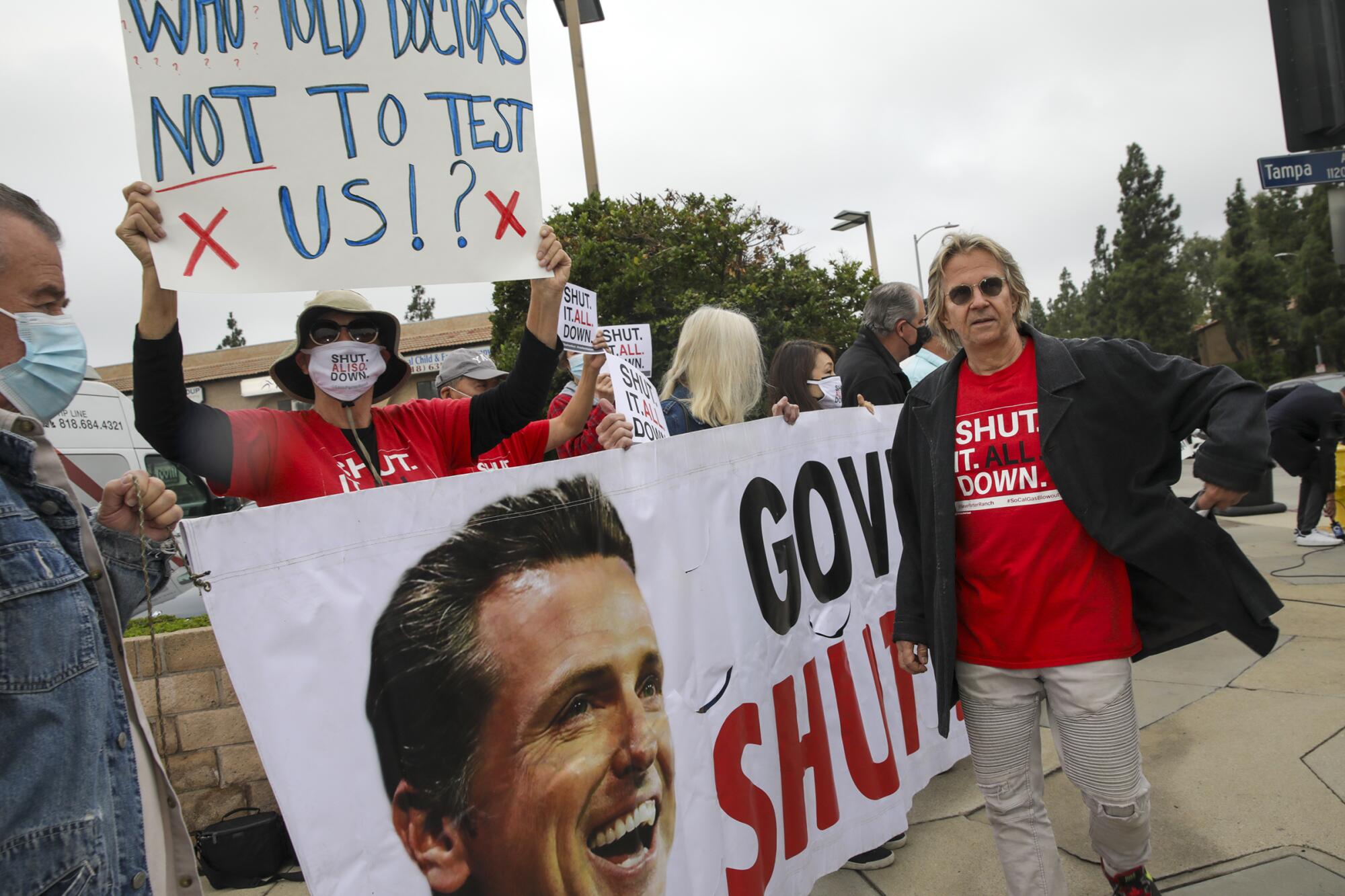 Un homme avec des lunettes, vêtu d'une veste sombre et d'une chemise rouge, se tient près de personnes tenant une pancarte et une bannière.