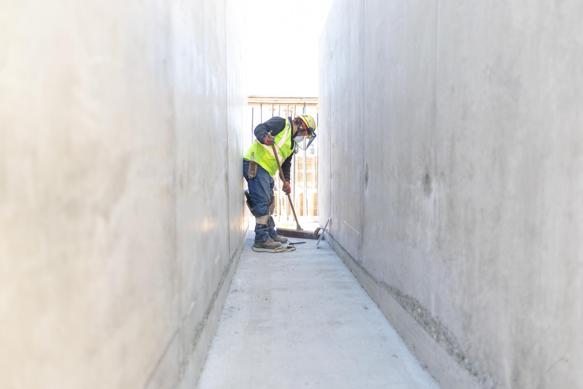 Une personne pousse un balai dans un passage étroit entre des murs en béton.
