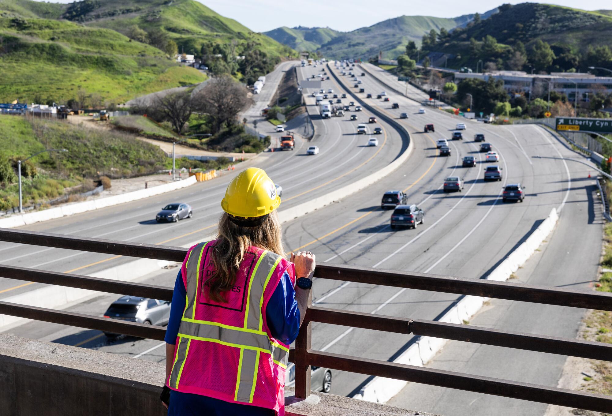 Une femme blonde aux cheveux longs, portant un casque jaune et un gilet de sécurité rose, regarde la circulation sur l'autoroute 101.
