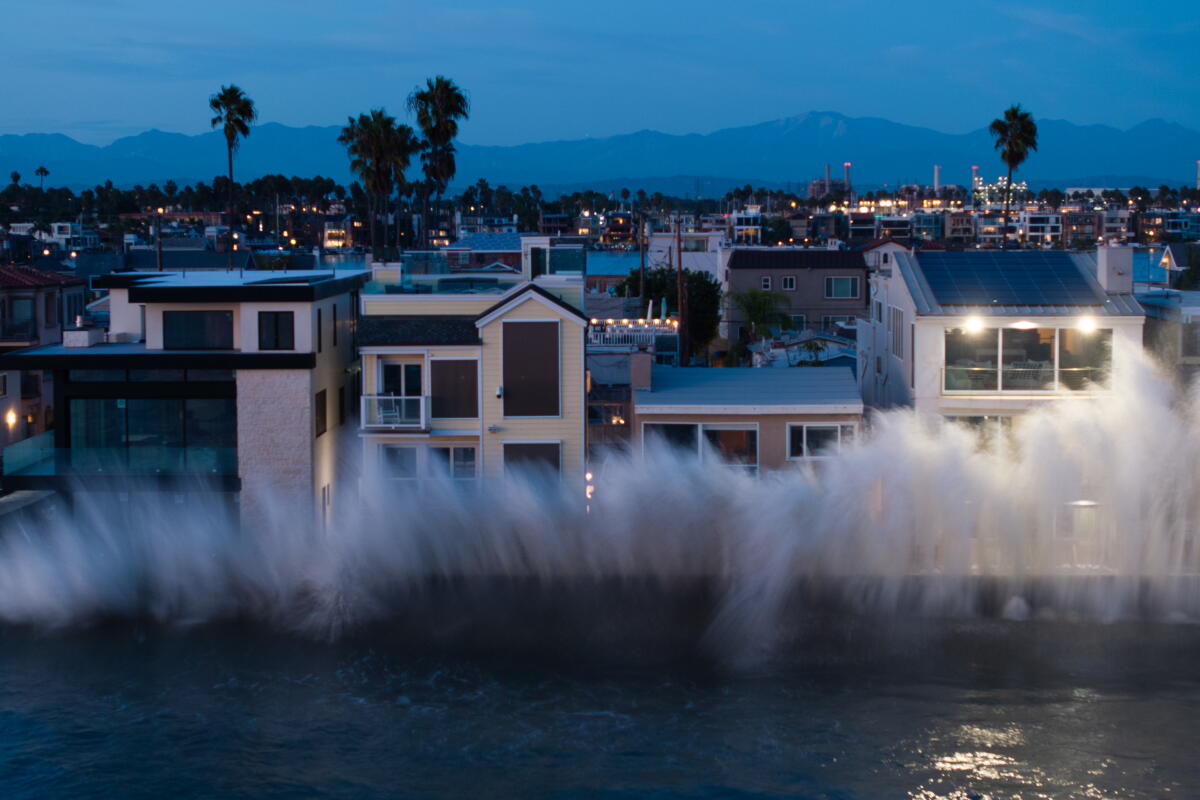 Une grande vague océanique frappe une rangée de maisons à plusieurs étages rapprochées à la tombée de la nuit.
