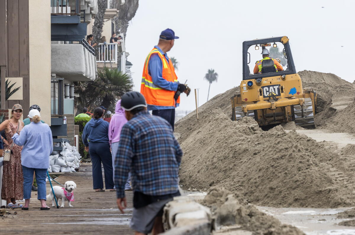 Les gens se rassemblent le long de la promenade alors que des machines lourdes construisent des bermes de sable sur la péninsule de Long Beach samedi.