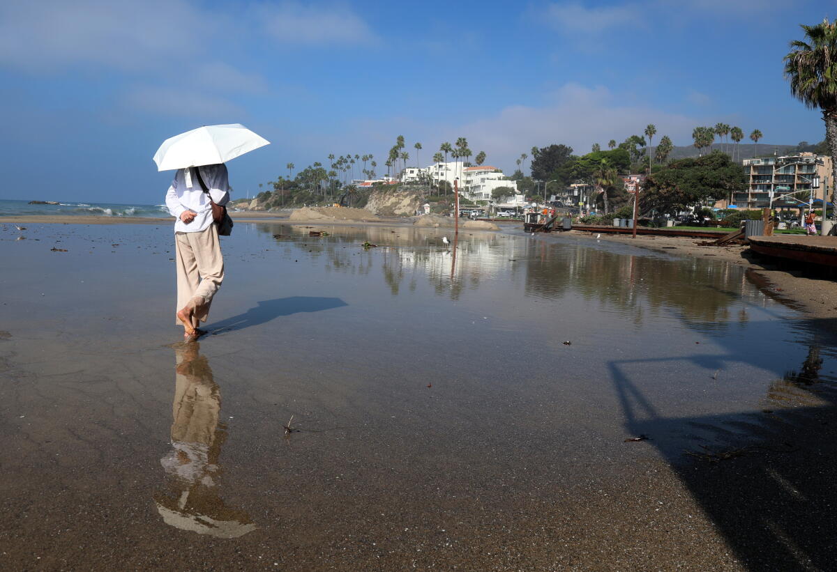 Une femme vêtue d'une tenue de couleur claire portant un parapluie de couleur claire se reflète dans l'eau de la plage.